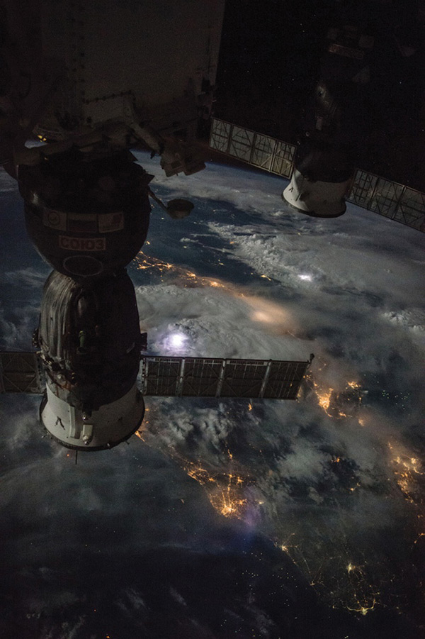 Far below the International Space Station, lightning flashes illuminate the clouds, as human activity is revealed by clusters of lights. Two Russian spacecraft visiting the station can be seen in the foreground.